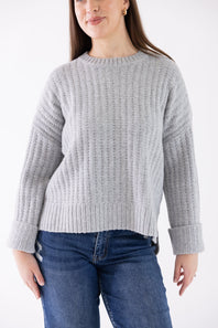 A woman wearing a light gray ribbed knit sweater and blue jeans, shown from the nose down against a plain white studio background.