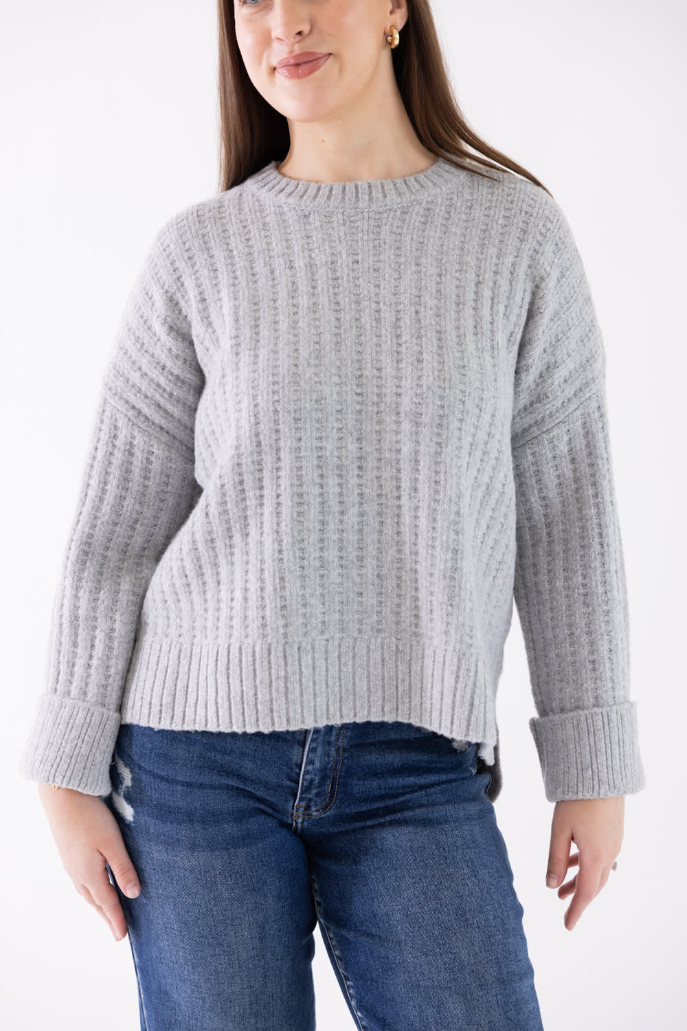 A woman wearing a light gray ribbed knit sweater and blue jeans, shown from the nose down against a plain white studio background.