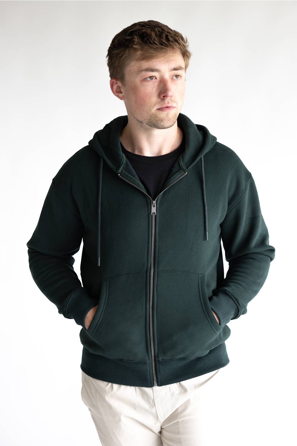 A man wearing a dark green zip-up hoodie over a black t-shirt and off-white trousers, standing against a plain white studio background.