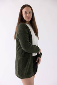 A woman modeling a dark olive green waffle-knit cardigan over a white top and black skirt, photographed against a plain white studio background.
