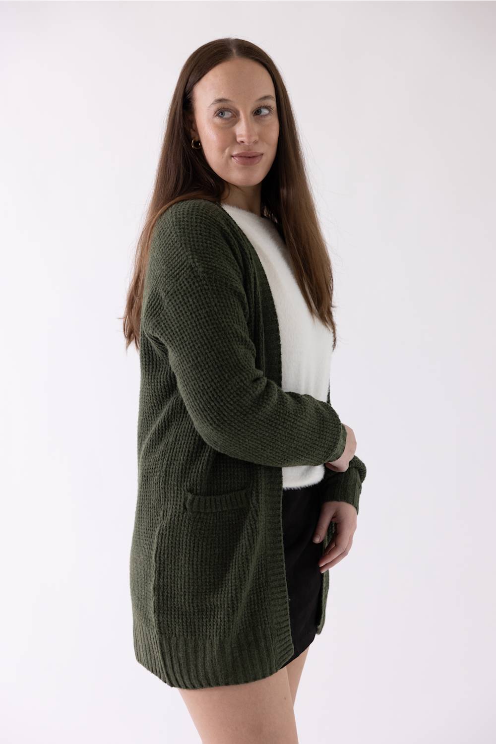 A woman modeling a dark olive green waffle-knit cardigan over a white top and black skirt, photographed against a plain white studio background.