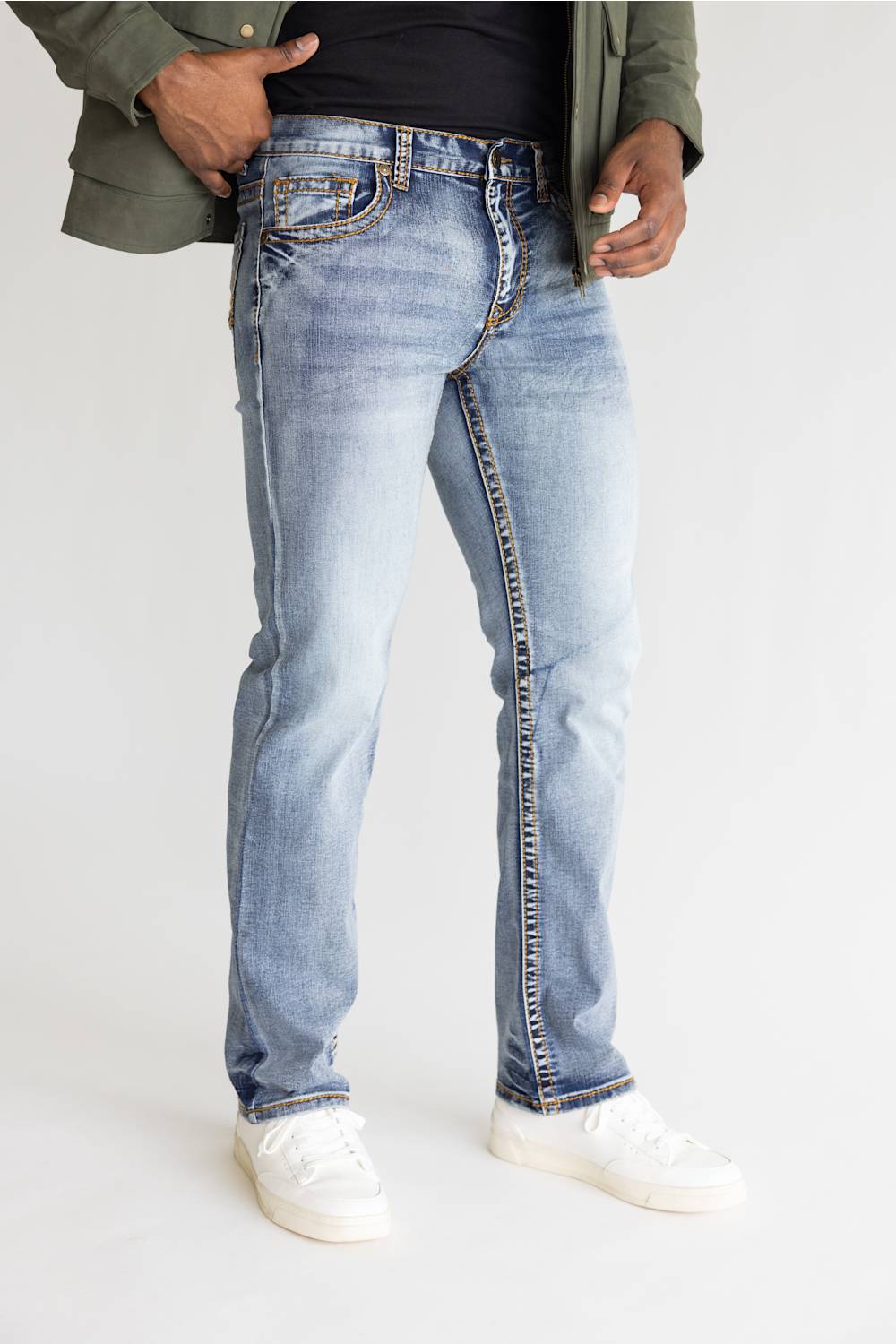 A person wearing light-wash blue denim jeans with a faded wash, styled with a black top, olive green jacket, and white sneakers against a plain white studio background.