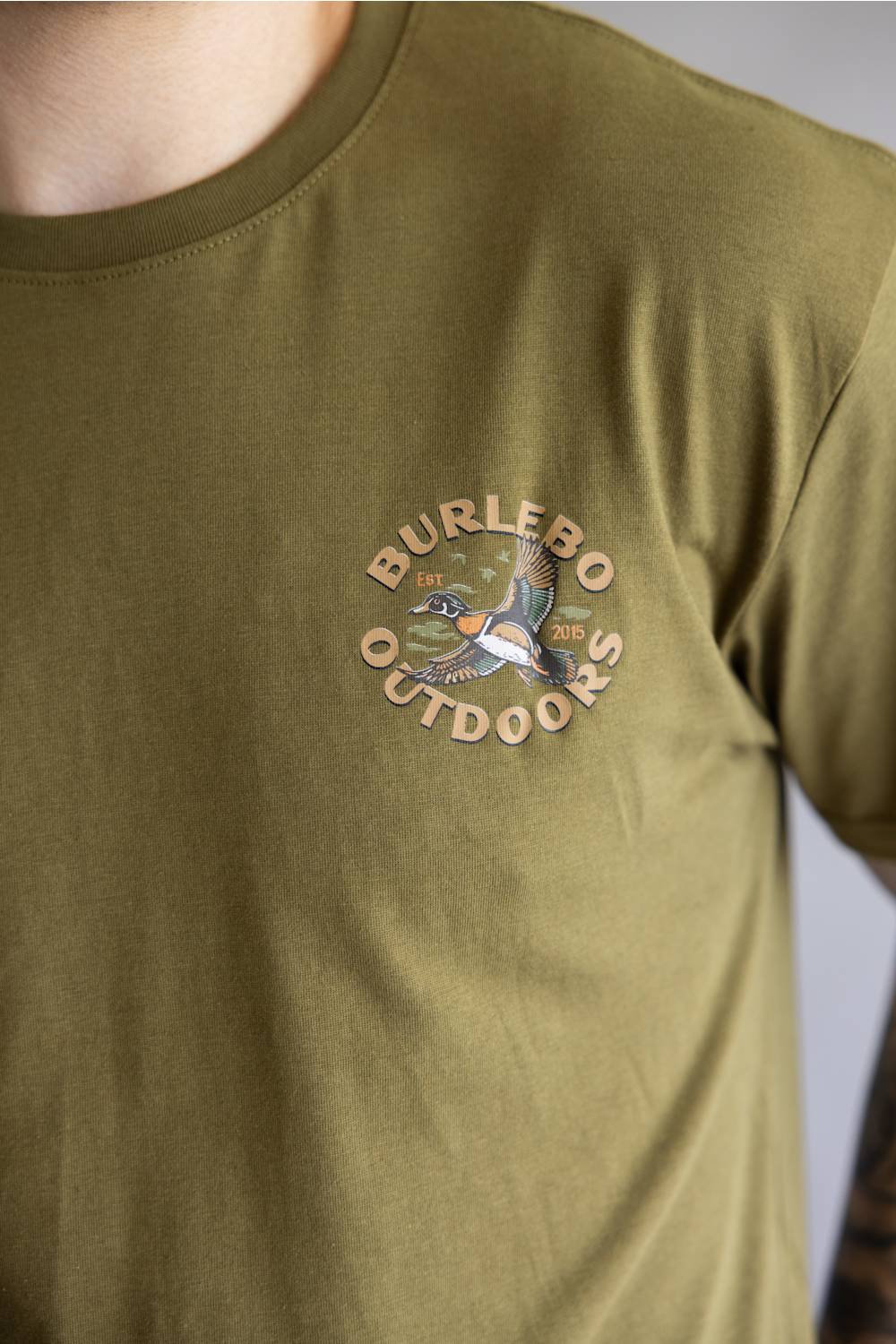 Close-up of a person wearing an olive green t-shirt with a circular 'BURLEBO OUTDOORS' duck logo printed on the left chest.
