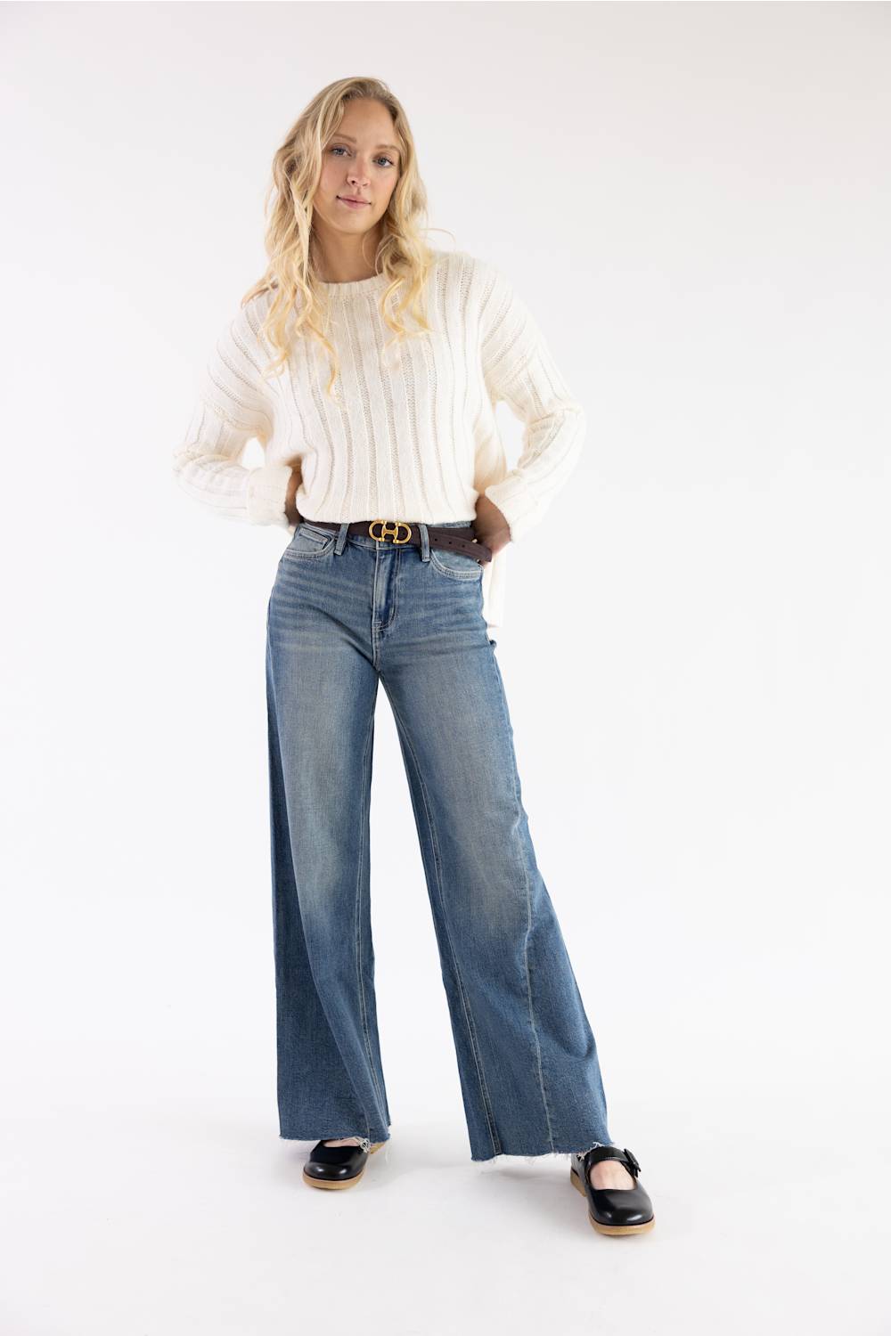 A woman modeling a cream ribbed knit sweater and wide-leg blue jeans with a brown belt, standing against a plain white studio background.