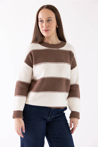 A woman wearing a brown and cream striped knit sweater with dark blue jeans, standing against a plain white studio background.