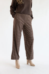 A person wearing brown wide-leg pants with thin beige horizontal stripes, paired with a dark brown sweater and beige heeled shoes, photographed against a plain white studio background.