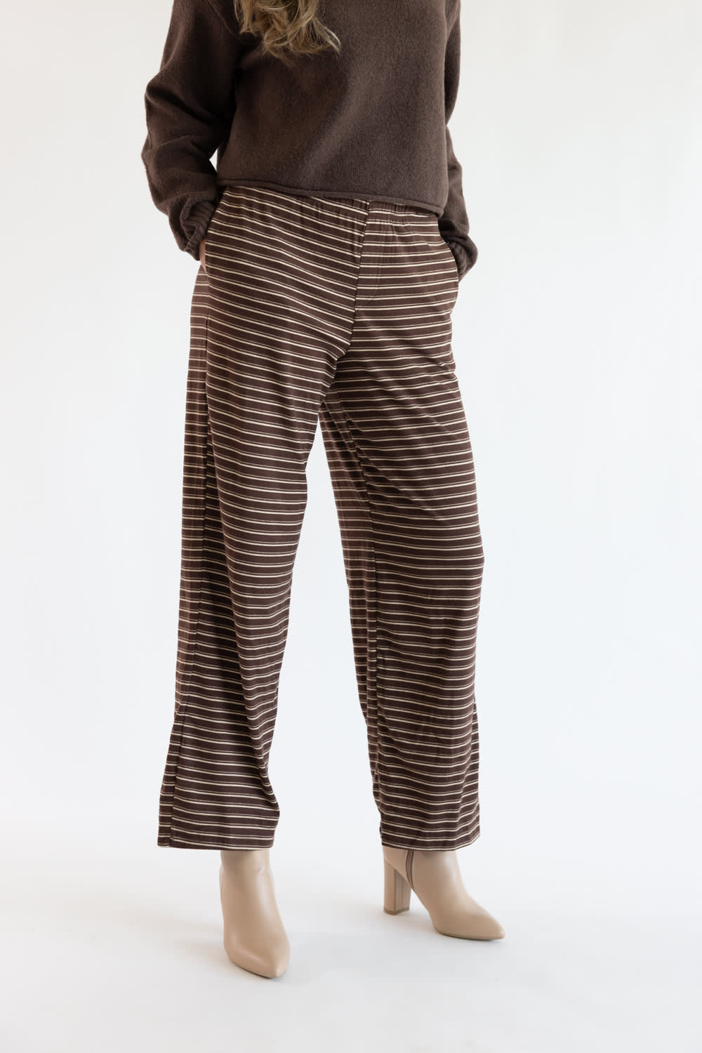 A person wearing brown wide-leg pants with thin beige horizontal stripes, paired with a dark brown sweater and beige heeled shoes, photographed against a plain white studio background.