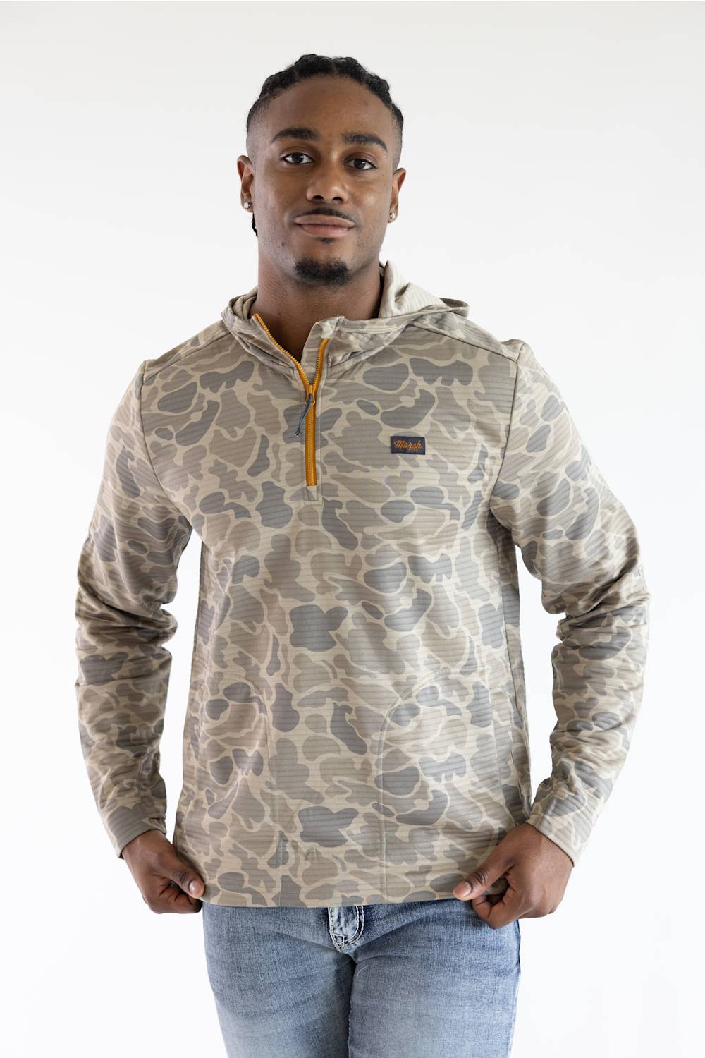 A man wearing a camouflage-patterned pullover hoodie with a quarter-zip collar and a small chest logo patch, paired with blue jeans, against a plain white studio background.