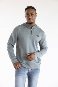 A man wearing a light gray ribbed quarter-zip pullover hoodie with a small brown logo patch on the chest, paired with light-wash blue jeans, photographed against a plain white studio background.