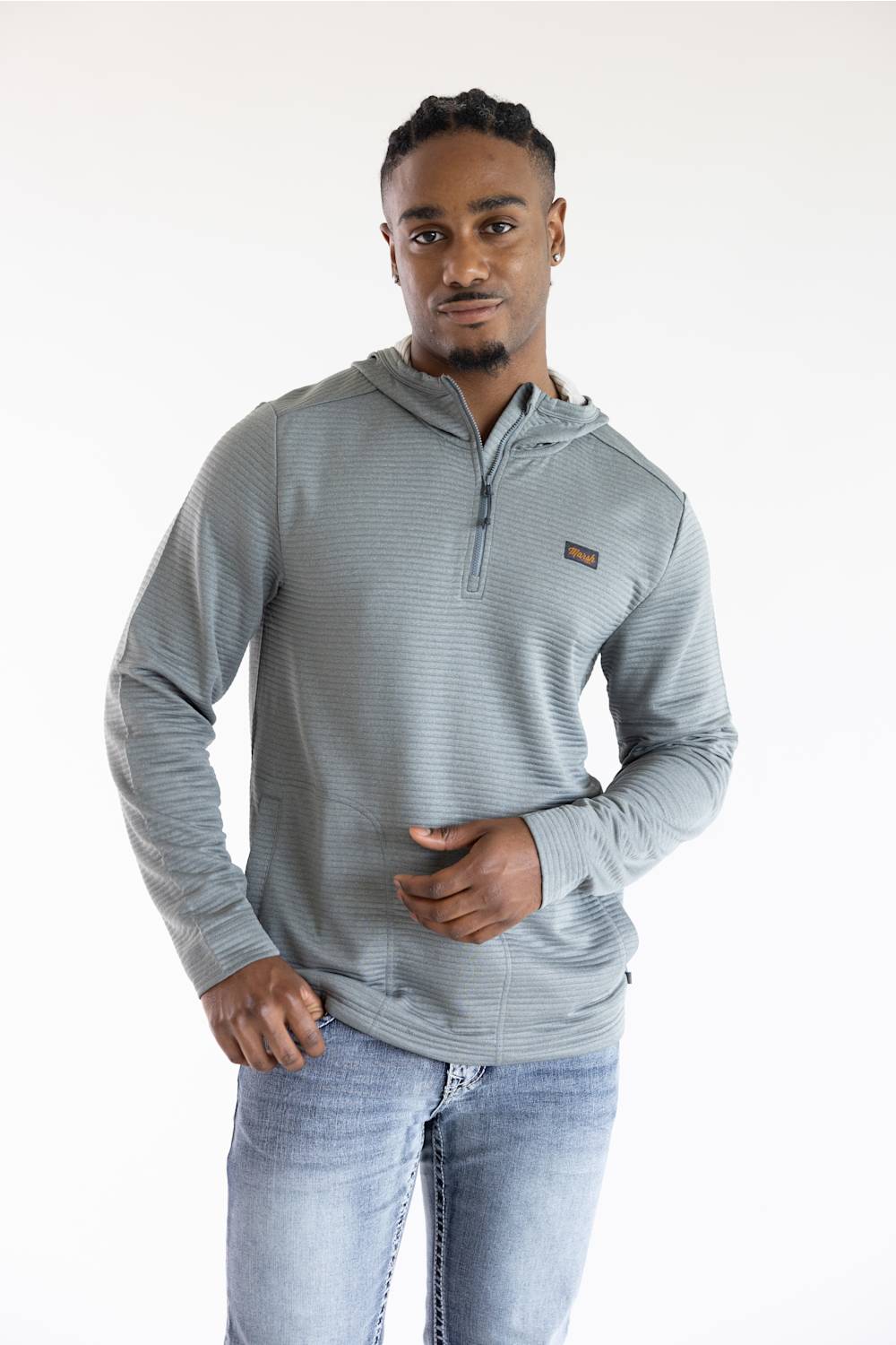 A man wearing a light gray ribbed quarter-zip pullover hoodie with a small brown logo patch on the chest, paired with light-wash blue jeans, photographed against a plain white studio background.
