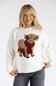 Blonde model wearing a cream crewneck sweater with an embroidered graphic of a fuzzy highland cow wearing a red and black plaid scarf, paired with dark grey jeans.