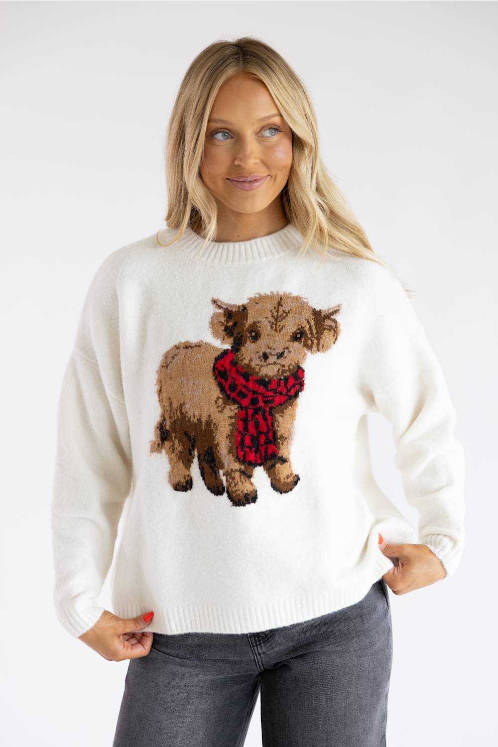 Blonde model wearing a cream crewneck sweater with an embroidered graphic of a fuzzy highland cow wearing a red and black plaid scarf, paired with dark grey jeans.