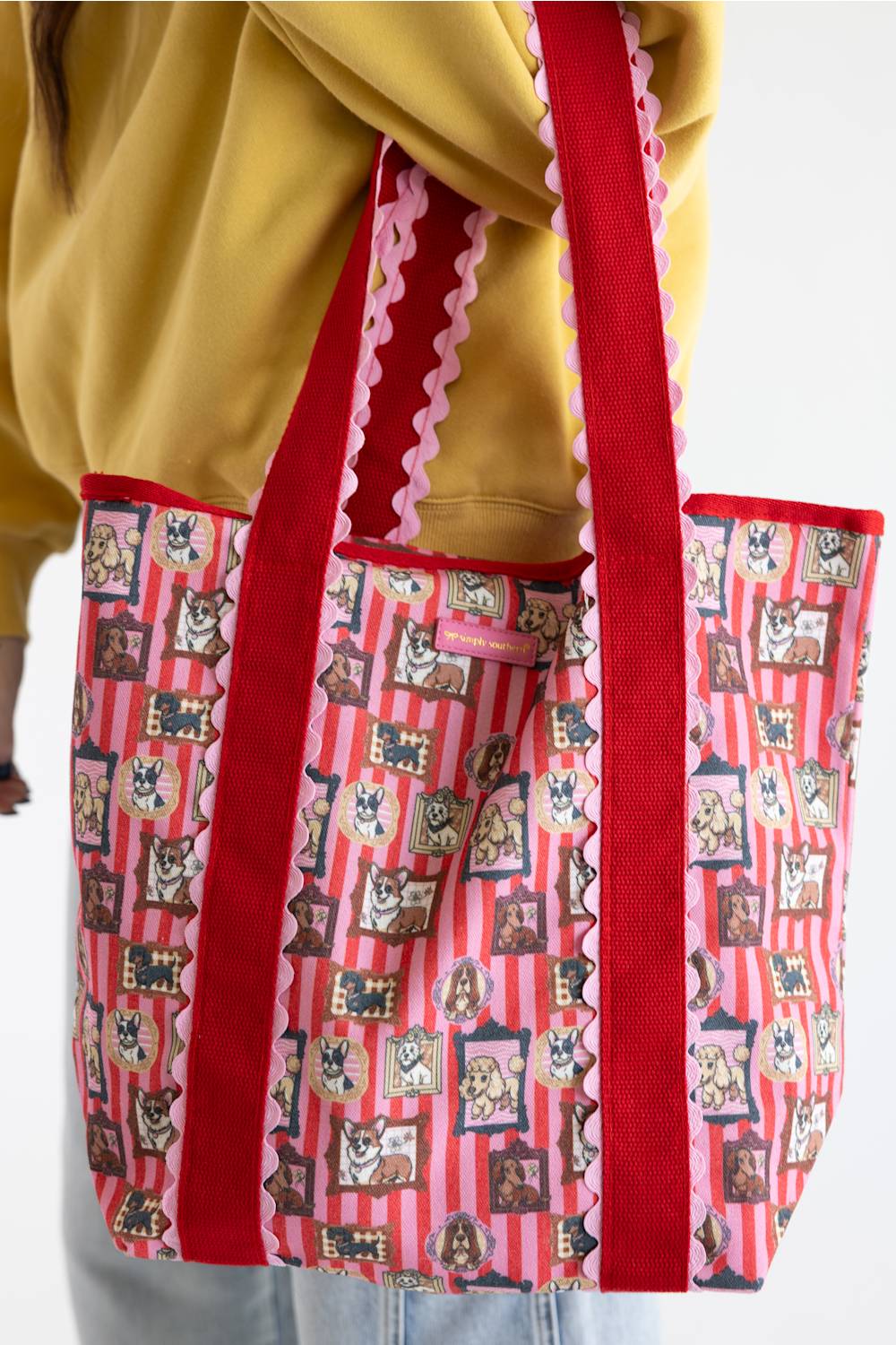 A red and pink patterned tote bag with dog illustrations, worn over the shoulder of a person wearing a yellow sweatshirt.