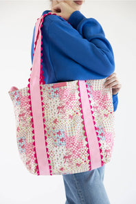 A person wearing a blue sweatshirt and jeans carries a large tote bag with a pink and multicolor floral print and pink striped handles against a plain white background.