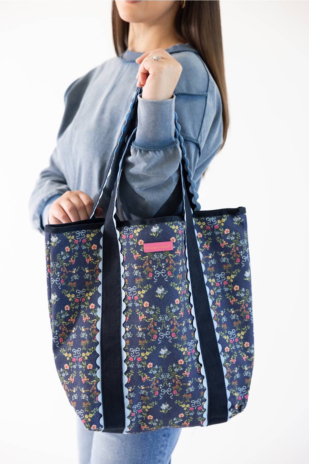 A woman wearing a blue sweatshirt holds up a navy tote bag with a colorful floral print and black straps against a plain white background.