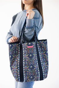 A woman wearing a blue sweatshirt holds up a navy tote bag with a colorful floral print and black straps against a plain white background.