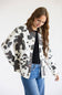 A woman wearing a cow-print jacket with brown faux-leather trim, a black top, and blue jeans, photographed against a plain white studio background.