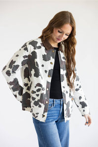 A woman wearing a cow-print jacket with brown faux-leather trim, a black top, and blue jeans, photographed against a plain white studio background.