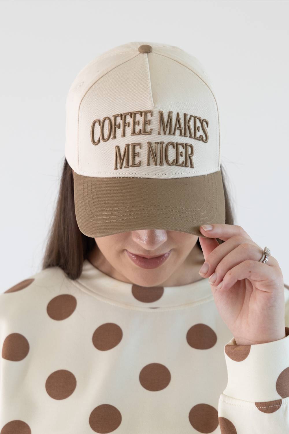 A woman wearing a cream and tan trucker hat with the embroidered text 'COFFEE MAKES ME NICER' in brown, paired with a cream long-sleeve top with brown polka dots, against a plain light gray background.