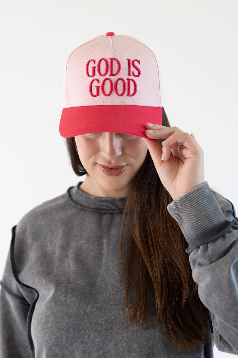A woman wearing a pink and red trucker hat with the words 'GOD IS GOOD' embroidered in red on the front, holding the brim with her hand against a plain white background.