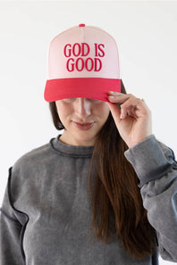 A woman wearing a pink and red trucker hat with the words 'GOD IS GOOD' embroidered in red on the front, holding the brim with her hand against a plain white background.