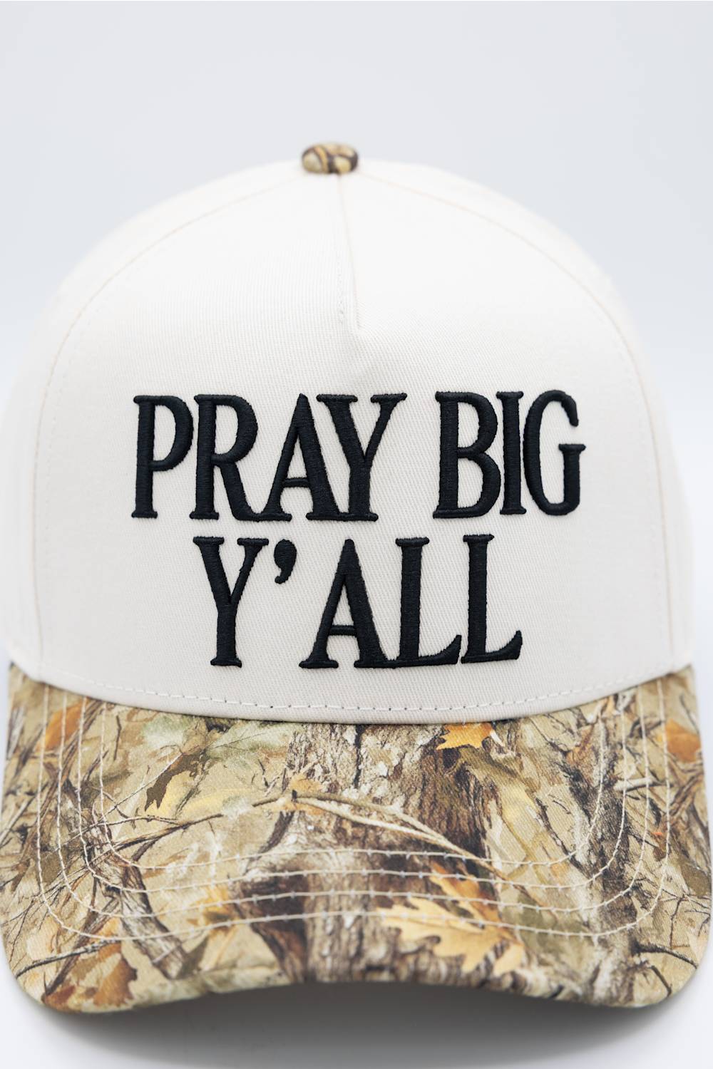 A baseball cap with an off-white crown and a camouflage-patterned brim. The front of the cap features the text 'PRAY BIG Y'ALL' embroidered in black.
