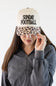 A woman wearing a cream-colored trucker hat with a leopard-print brim and the words 'SUNDAY FOOTBALL' printed in black on the front. She is wearing a dark gray quarter-zip sweatshirt against a plain white background.