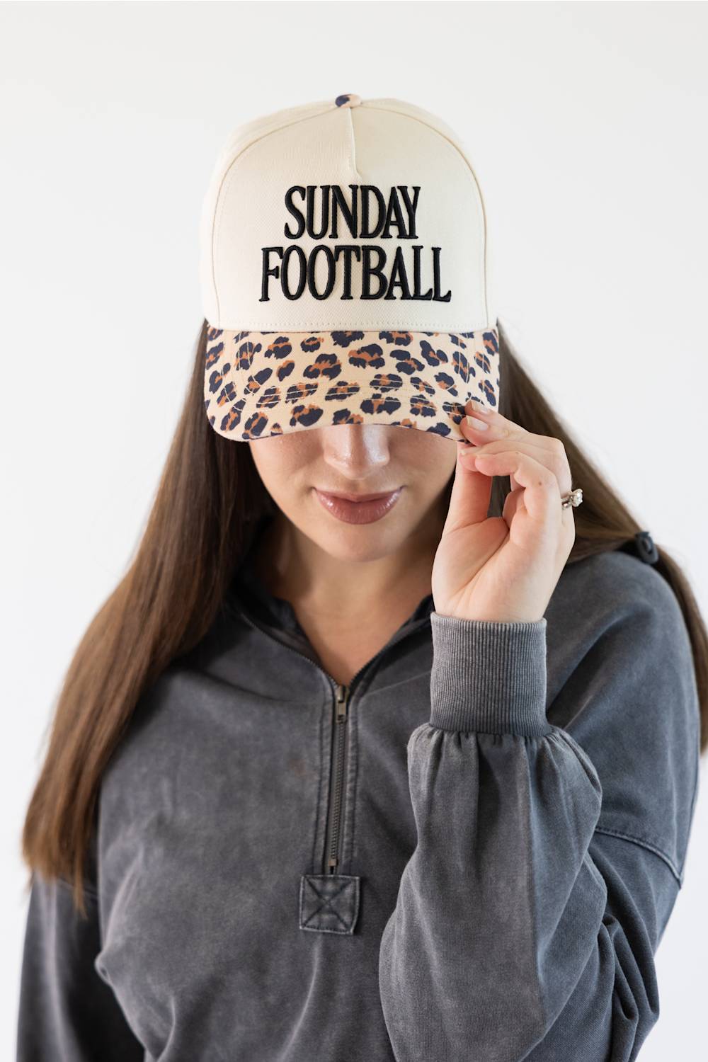 A woman wearing a cream-colored trucker hat with a leopard-print brim and the words 'SUNDAY FOOTBALL' printed in black on the front. She is wearing a dark gray quarter-zip sweatshirt against a plain white background.