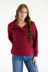 A woman modeling a burgundy half-zip pullover sweatshirt with side drawstring details, paired with blue jeans, against a plain white studio background.
