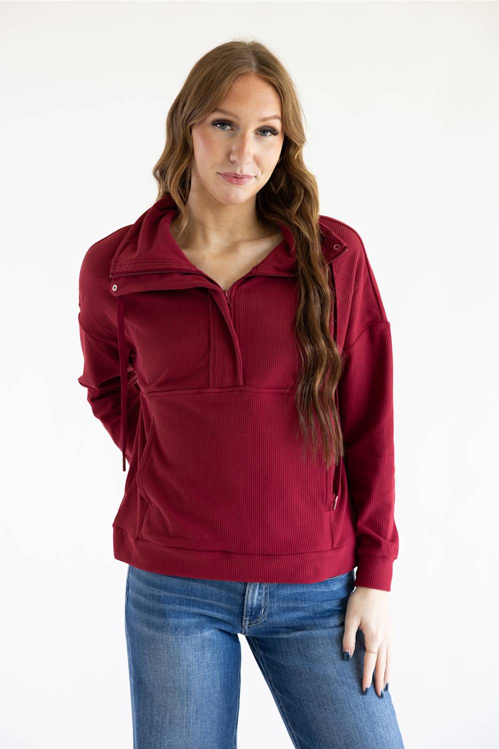 A woman modeling a burgundy half-zip pullover sweatshirt with side drawstring details, paired with blue jeans, against a plain white studio background.