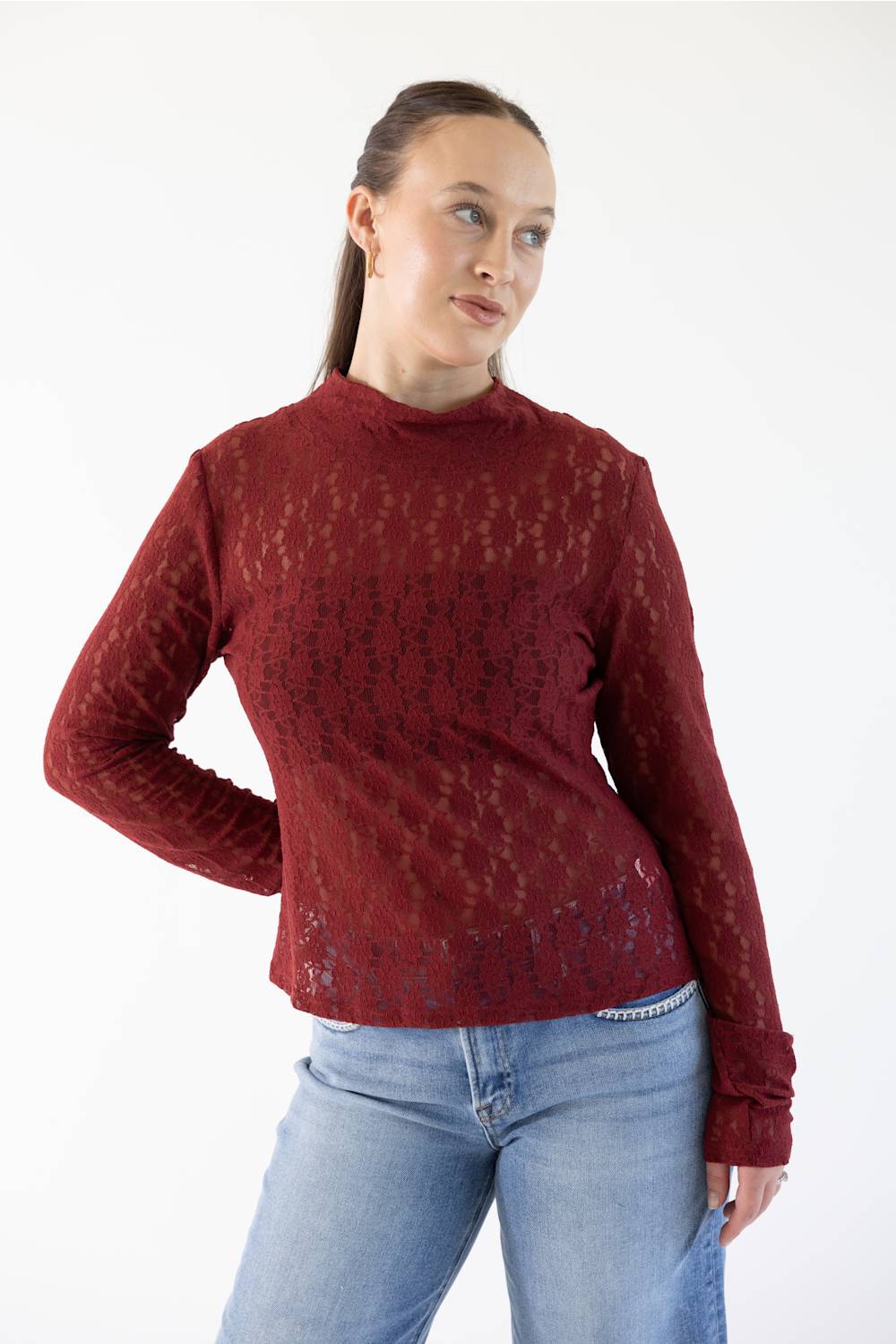 A woman modeling a long-sleeve, mock-neck knit top in a deep burgundy red, paired with light-wash blue jeans, against a plain white studio background.