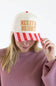 Model touching the brim of the Simply Southern Merry & Bright Holiday Hat for Women in Cream/Stripe, featuring yellow and red striped embroidered text reading "MERRY & BRIGHT" on the cream front panel and a red and pink striped visor, paired with a dusty pink long-sleeve top.