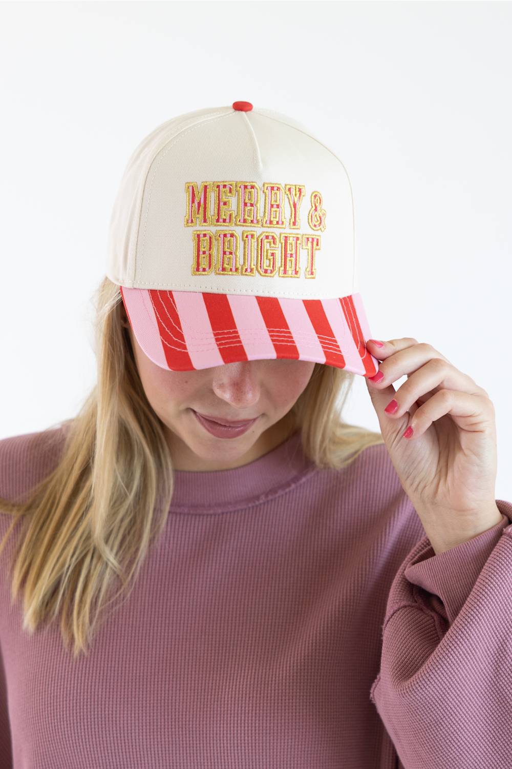 Model touching the brim of the Simply Southern Merry & Bright Holiday Hat for Women in Cream/Stripe, featuring yellow and red striped embroidered text reading "MERRY & BRIGHT" on the cream front panel and a red and pink striped visor, paired with a dusty pink long-sleeve top.