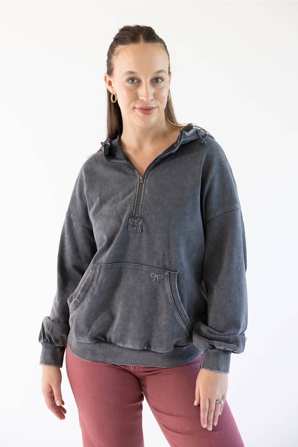 A woman modeling a dark gray washed half-zip hoodie with a drawstring hood and kangaroo pocket, paired with dusty rose pants, against a plain white studio background.