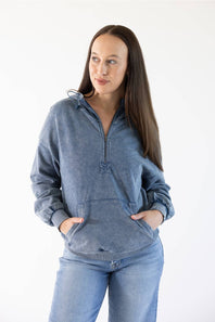 A woman modeling a washed slate blue half-zip pullover sweatshirt with a small cross detail at the center of the chest, paired with light blue jeans, against a plain white background.