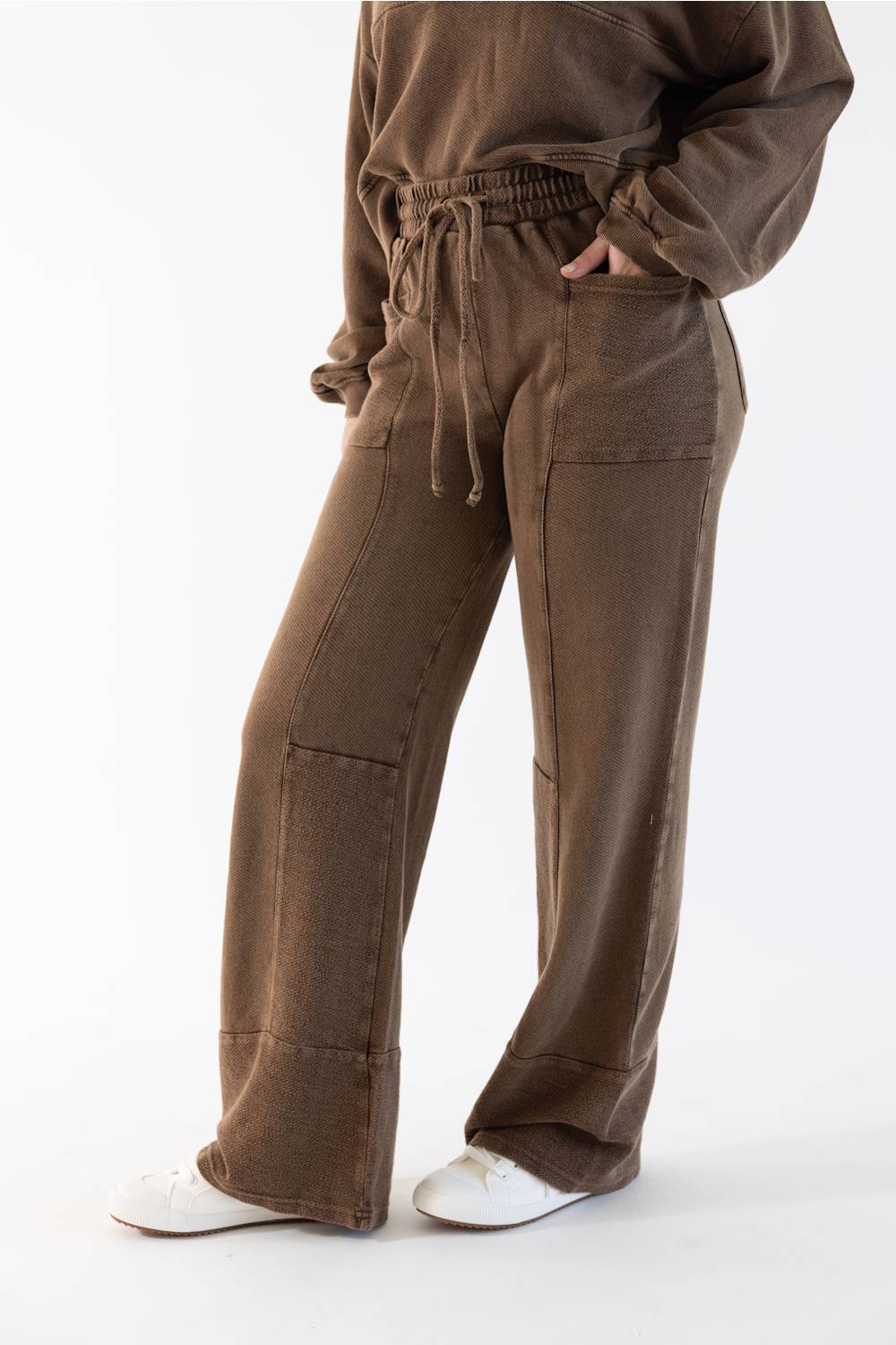 A person wearing brown wide-leg drawstring pants with a matching sweatshirt and white sneakers, shown from the chest down against a plain white studio background.