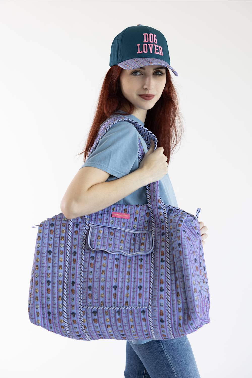 A woman wearing a blue denim t-shirt, jeans, and a navy blue baseball cap with 'DOG LOVER' embroidered in pink, carrying a large lavender and purple patterned tote bag over her shoulder, set against a plain white background.