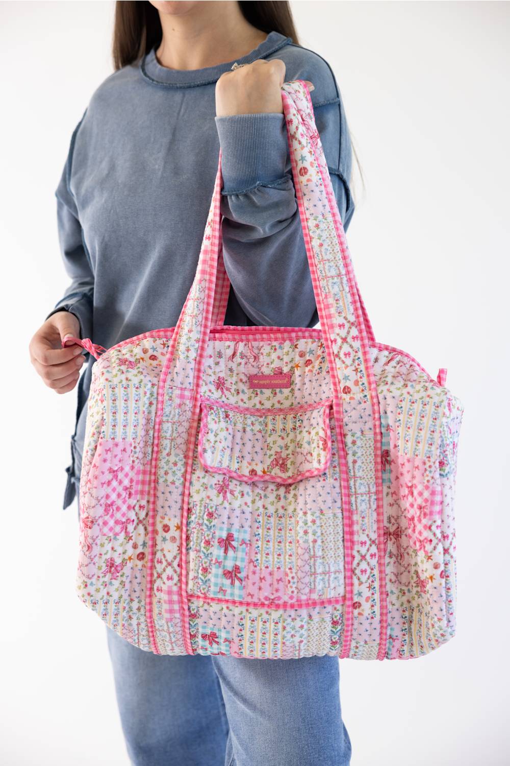 A person wearing a gray sweatshirt and blue jeans holds up a large quilted duffel bag with a pink and multicolor patchwork pattern. The bag features multiple pockets and long shoulder straps, shown against a plain white background.