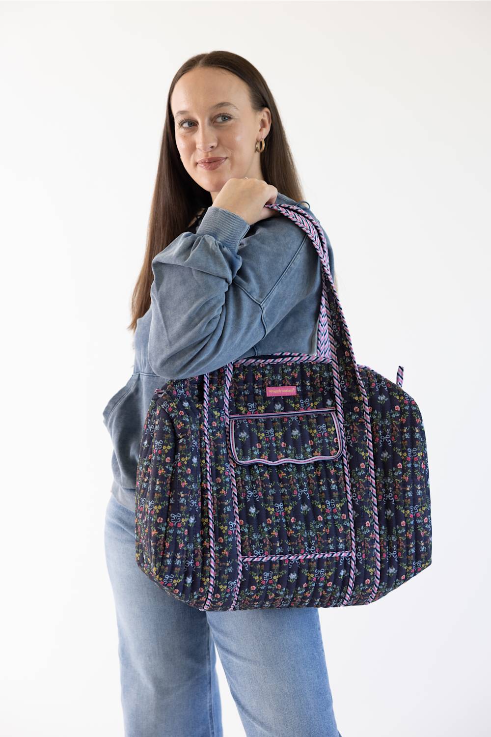 A woman wearing a blue denim jacket and jeans carries a large woven tote bag with a multicolor floral pattern and purple trim against a plain white background.