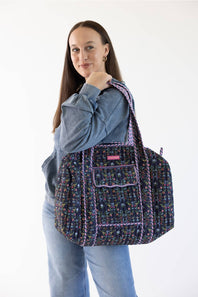 A woman wearing a blue denim jacket and jeans carries a large woven tote bag with a multicolor floral pattern and purple trim against a plain white background.