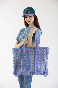 A woman wearing a blue 'DOG LOVER' cap, a gray t-shirt, and jeans holds a large purple patterned woven tote bag with fringe details against a plain white background.