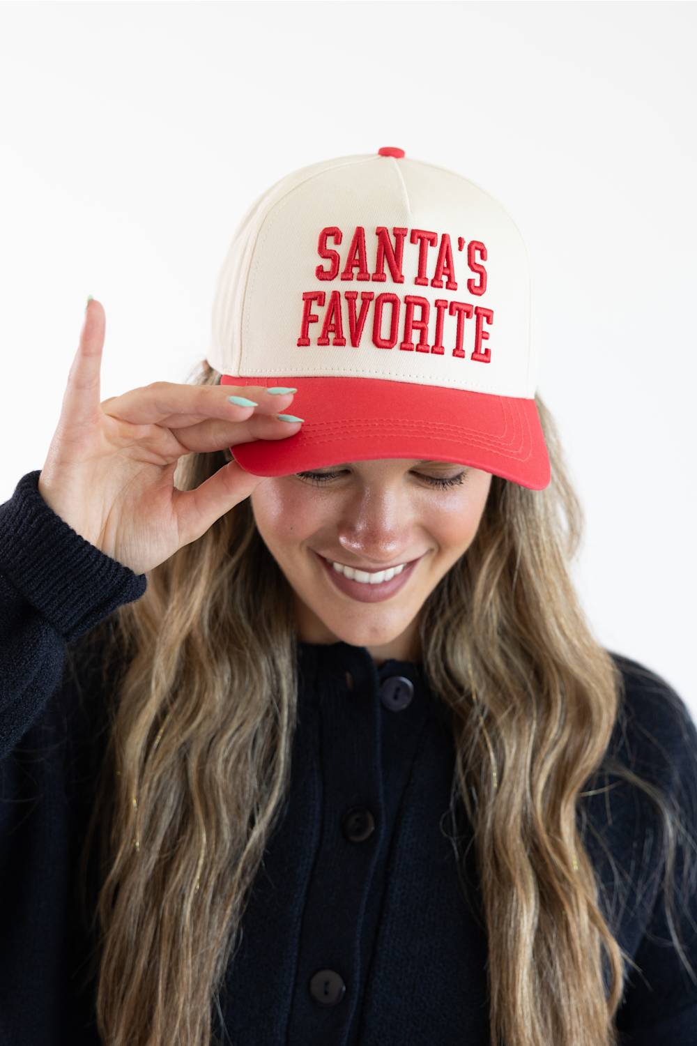 Model holding the brim of the Simply Southern Santa’s Favorite Holiday Hat for Women in Cream/Red, featuring red embroidered collegiate-style lettering reading “SANTA’S FAVORITE” on the cream front panel and a solid red visor, paired with a dark button-down cardigan.