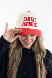 Model holding the brim of the Simply Southern Santa’s Favorite Holiday Hat for Women in Cream/Red, featuring red embroidered collegiate-style lettering reading “SANTA’S FAVORITE” on the cream front panel and a solid red visor, paired with a dark button-down cardigan.
