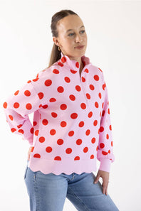 A woman modeling a pink long-sleeve quarter-zip pullover sweatshirt with a red polka dot pattern and scalloped edge, paired with blue jeans, against a plain white studio background.
