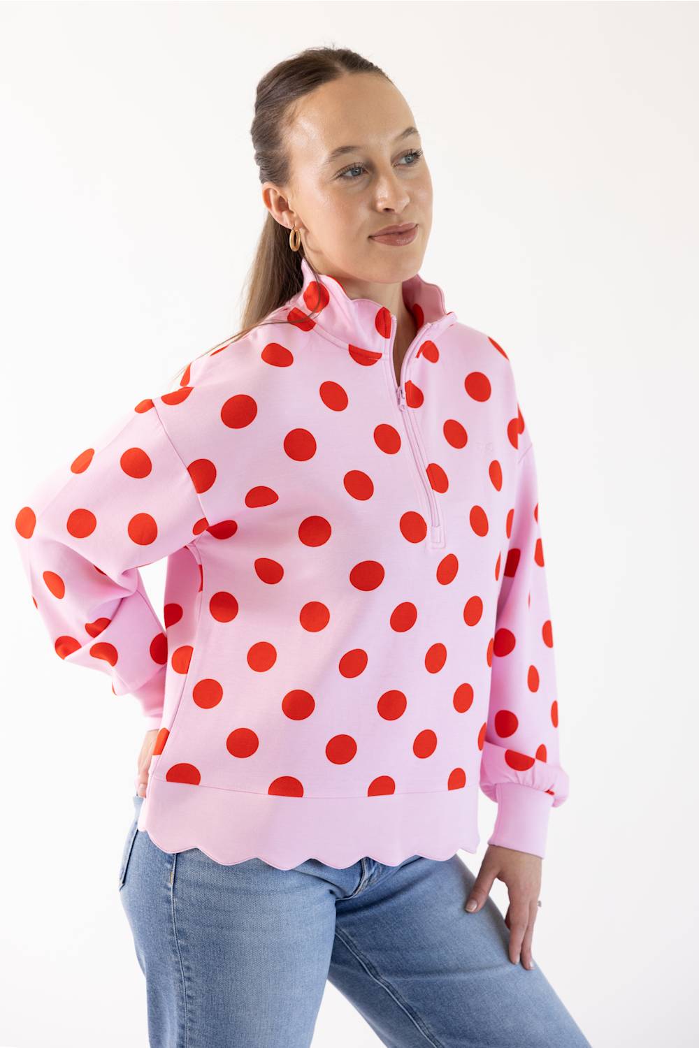 A woman modeling a pink long-sleeve quarter-zip pullover sweatshirt with a red polka dot pattern and scalloped edge, paired with blue jeans, against a plain white studio background.