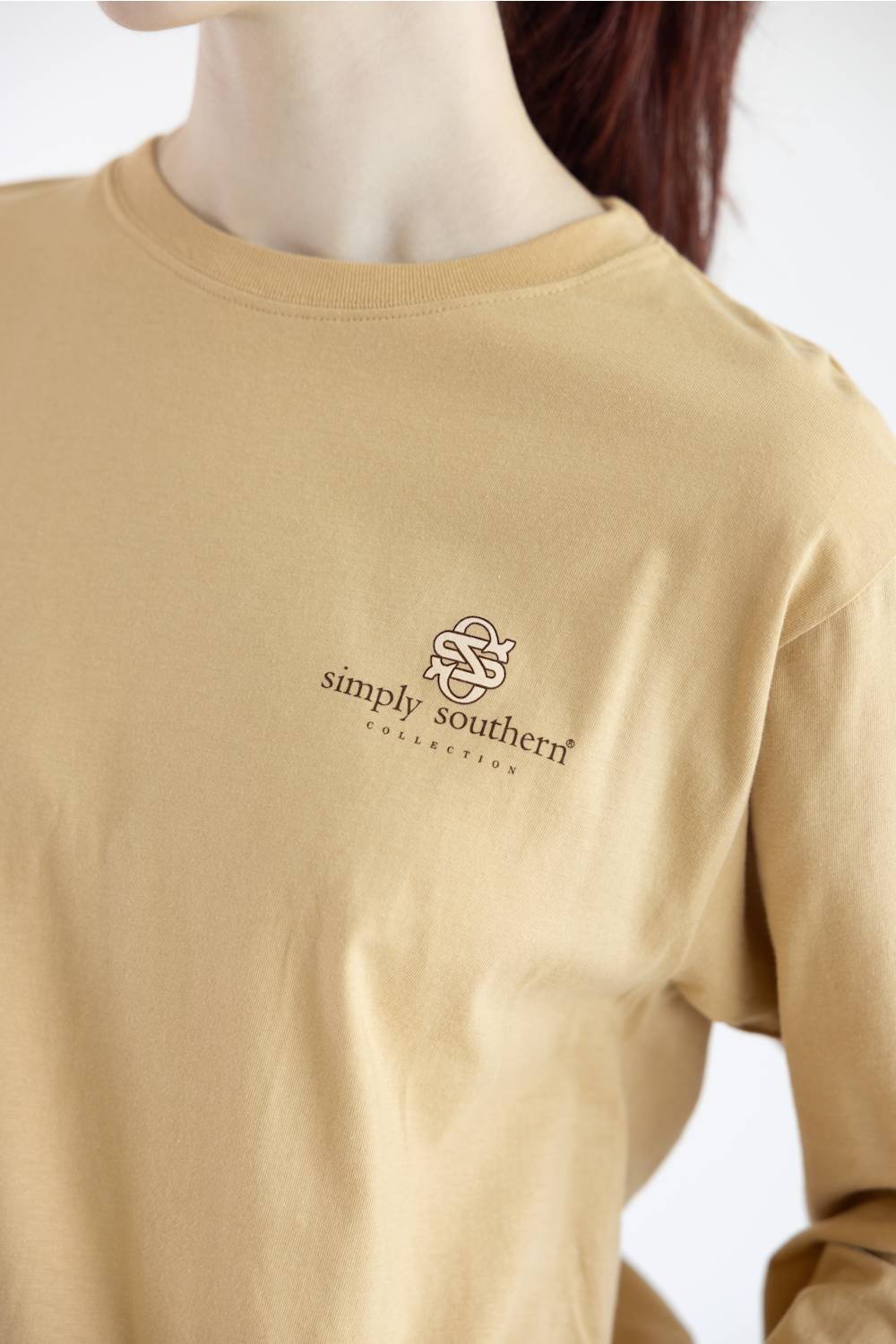 A close-up of a person wearing a tan long-sleeve crewneck shirt with a small embroidered 'simply southern' logo on the left chest, set against a plain white background.