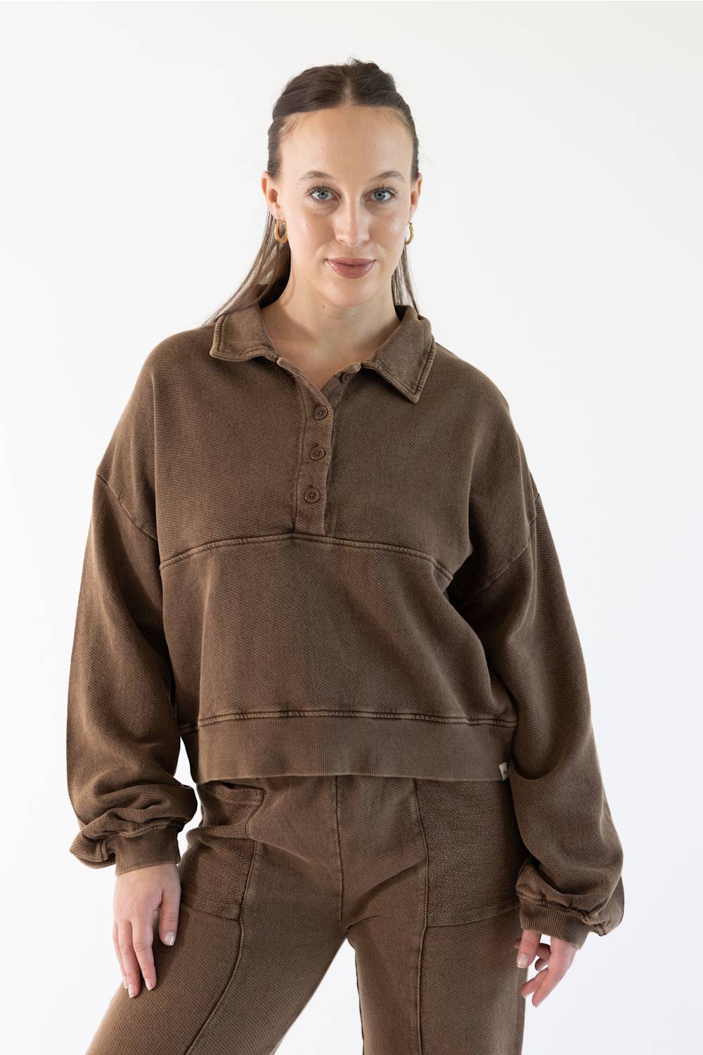 A woman modeling a brown cropped long-sleeve polo sweatshirt with matching brown pants against a plain white studio background.