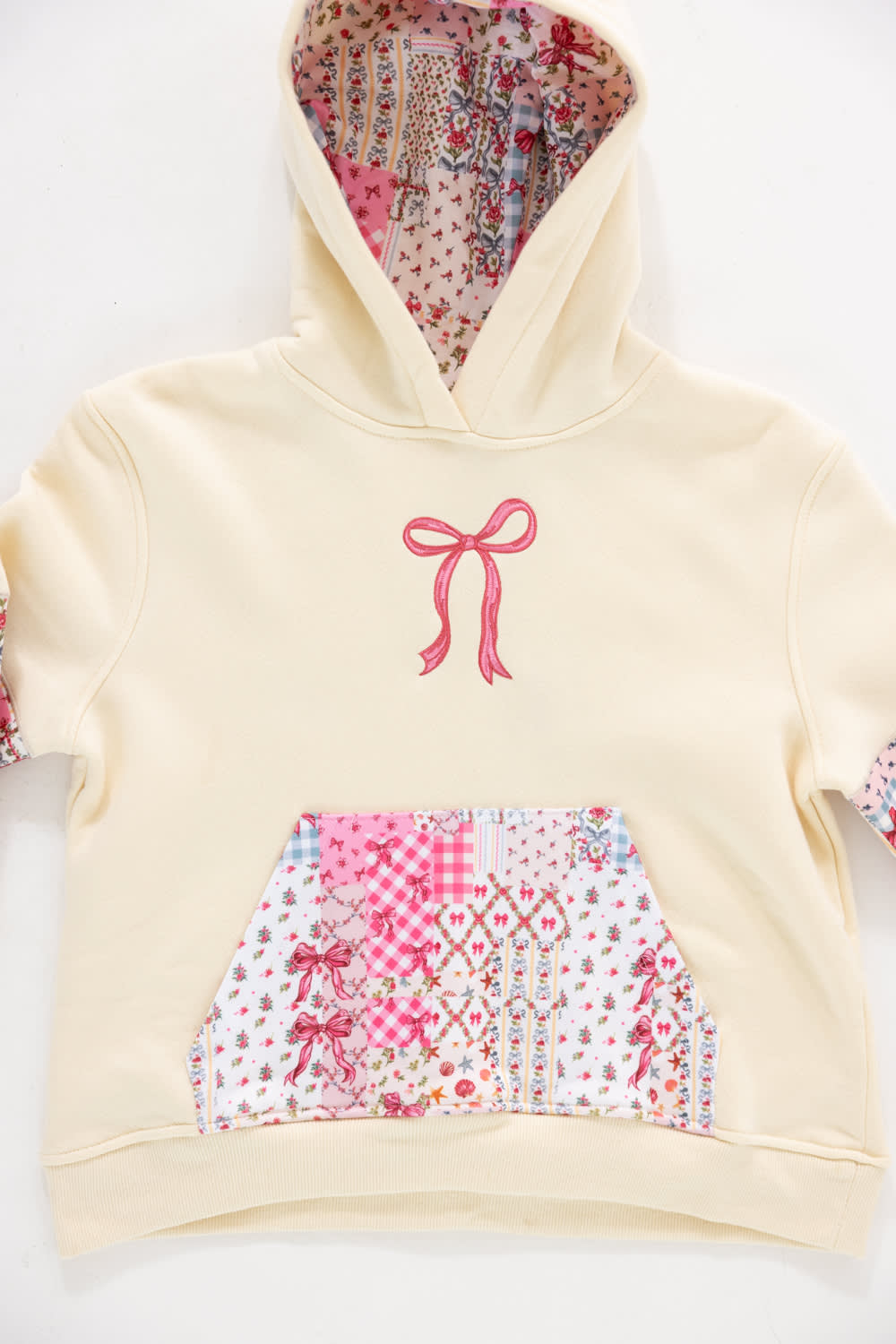 Close-up front view of a girls' cream-colored pullover hoodie showing an embroidered pink ribbon bow on the chest, a patchwork floral and gingham kangaroo pocket, and a floral-patterned lined hood against a plain white background.