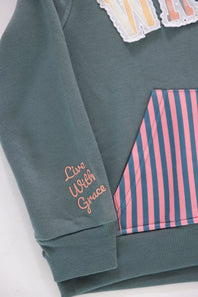 Close-up shot of the sleeve cuff on a girls' gray pullover hoodie featuring pink embroidered cursive text reading “Live With Grace”, with a portion of the pink and navy striped kangaroo pocket and patterned applique chest lettering visible.