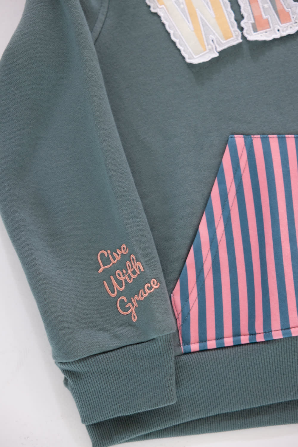 Close-up shot of the sleeve cuff on a girls' gray pullover hoodie featuring pink embroidered cursive text reading “Live With Grace”, with a portion of the pink and navy striped kangaroo pocket and patterned applique chest lettering visible.